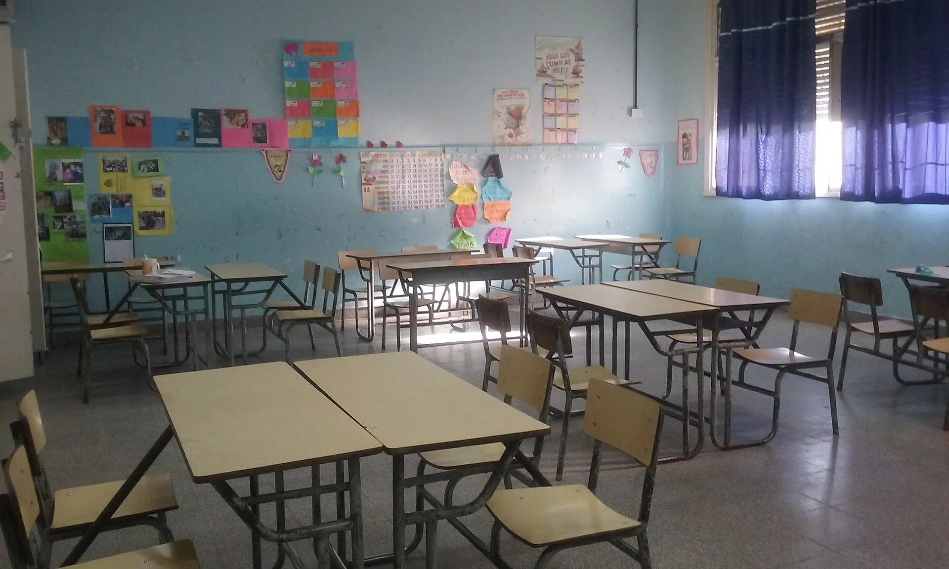An empty classroom with rows of wooden desks and chairs and colorful student work pinned to the walls.