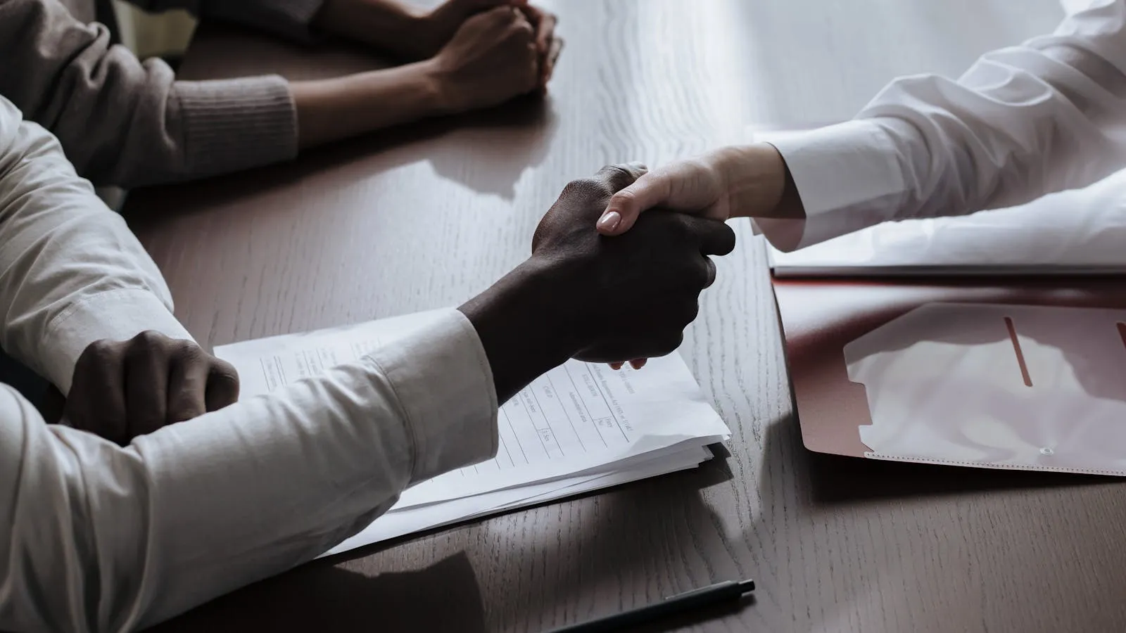 Two people in business attire extending hands for a formal handshake over a table with documents