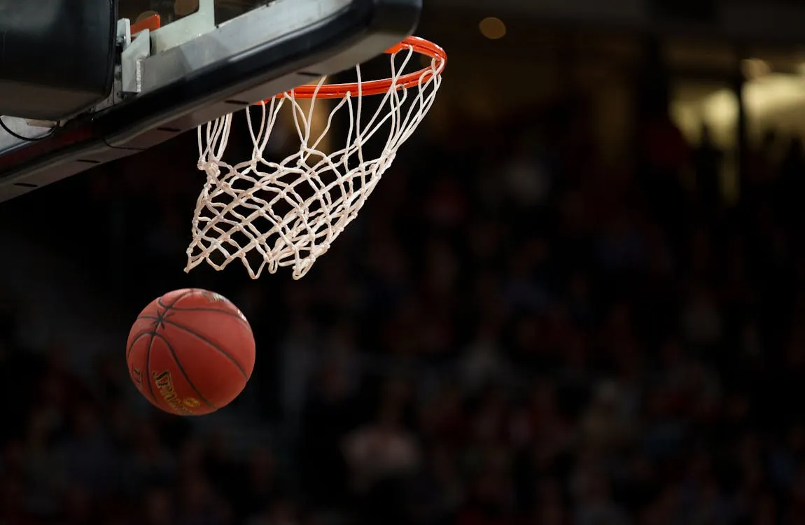 An orange basketball dropping through a white net on an indoor basketball hoop, with a blurred crowd visible in the background.