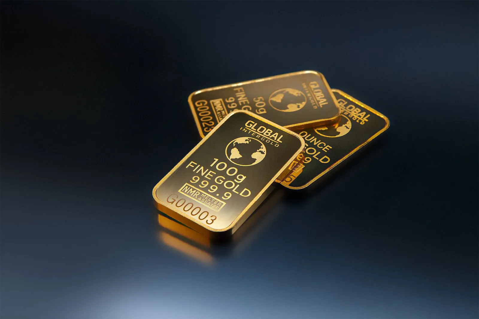 Three gold bars arranged on a dark surface, showing stamped markings and reflective surfaces