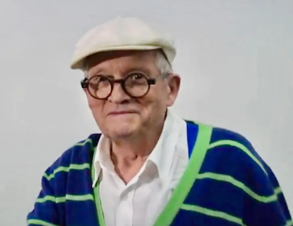 David Hockney, an older man in glasses and a cap, photographed at a public art event.