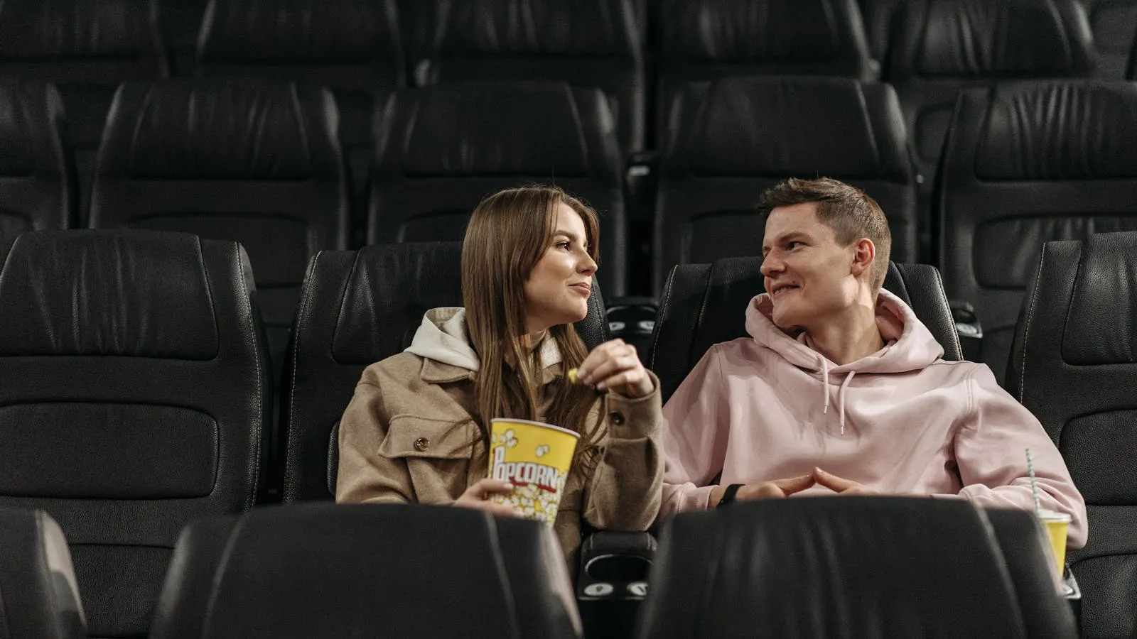 A couple seated in a darkened movie theater with a bucket of popcorn between them, looking toward the screen