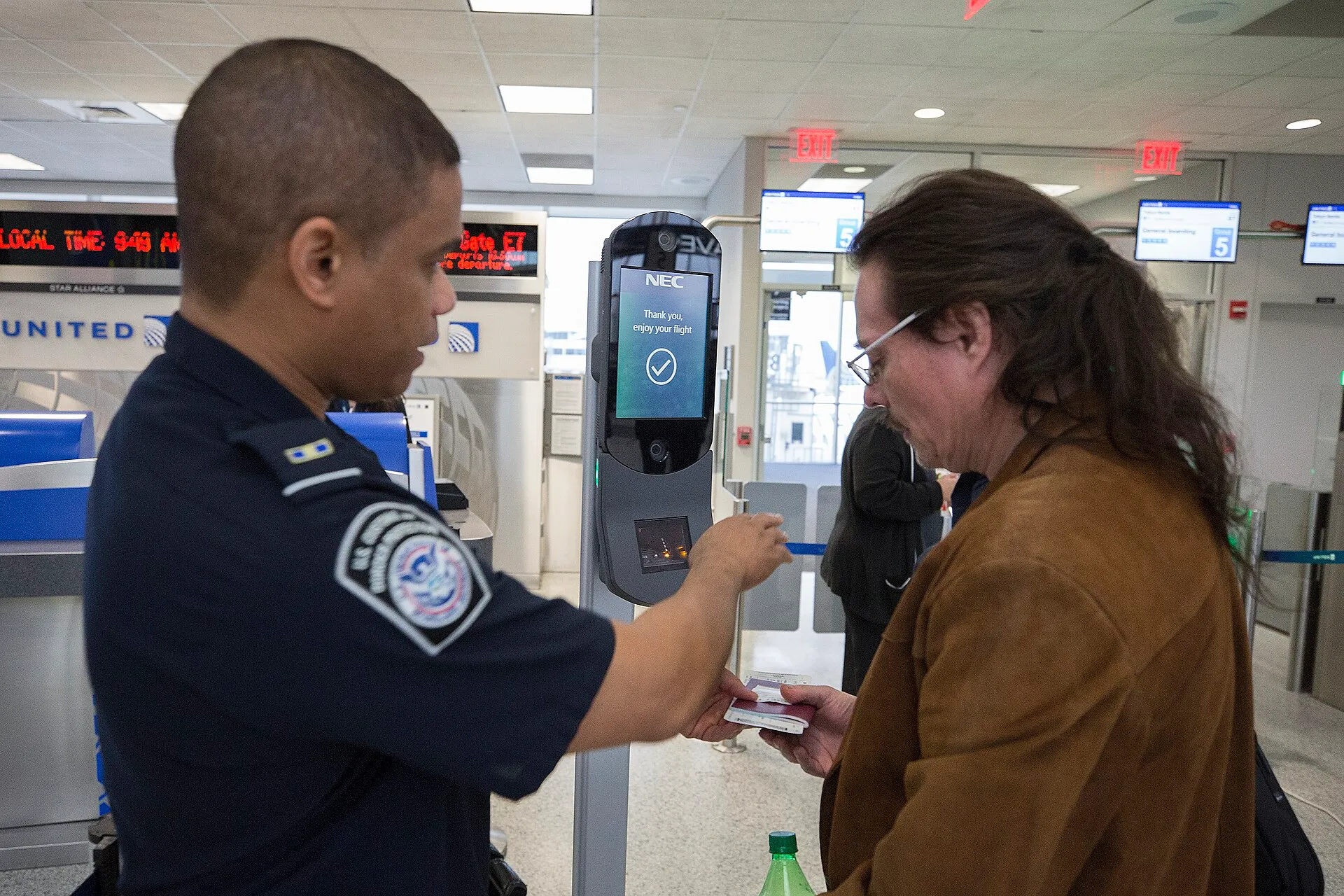 A Customs and Border Protection officer operates a facial-recognition kiosk as a traveler faces the camera at an airport gate.