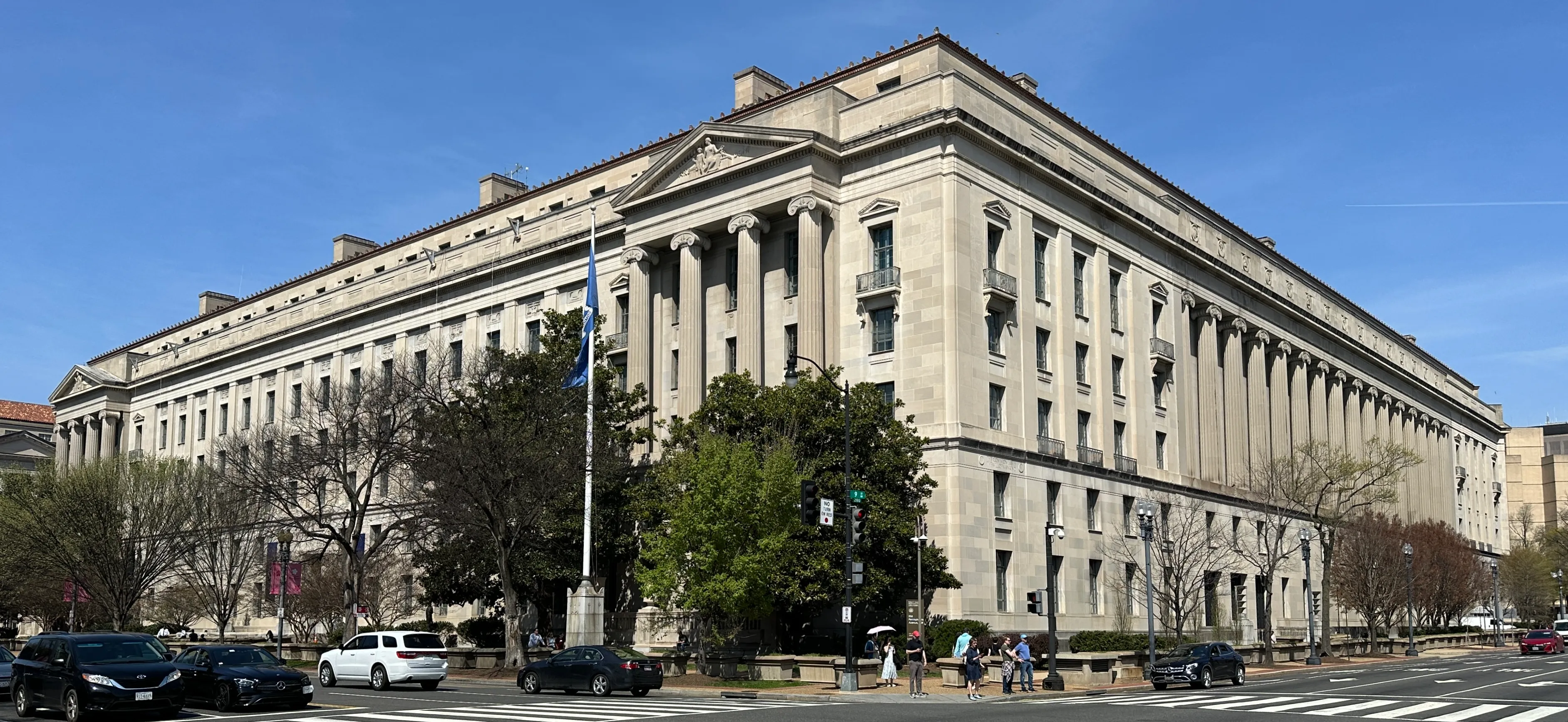The Robert F. Kennedy Department of Justice Building, a large neoclassical limestone structure with a colonnade, seen from a street corner under a clear blue sky.