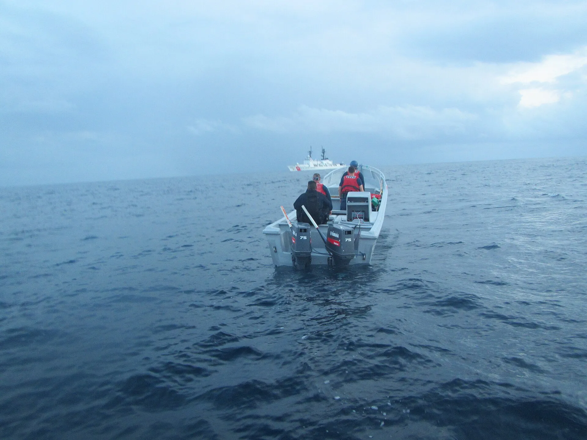A small open boat with twin outboard motors sits on the open ocean as a U.S. Coast Guard cutter waits in the distance under an overcast sky.