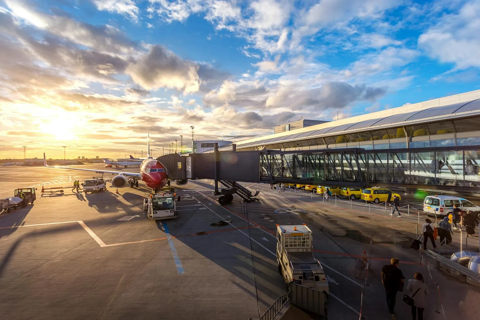 A commercial airplane parked at an airport gate at sunset, with ground crew vehicles and terminal building visible