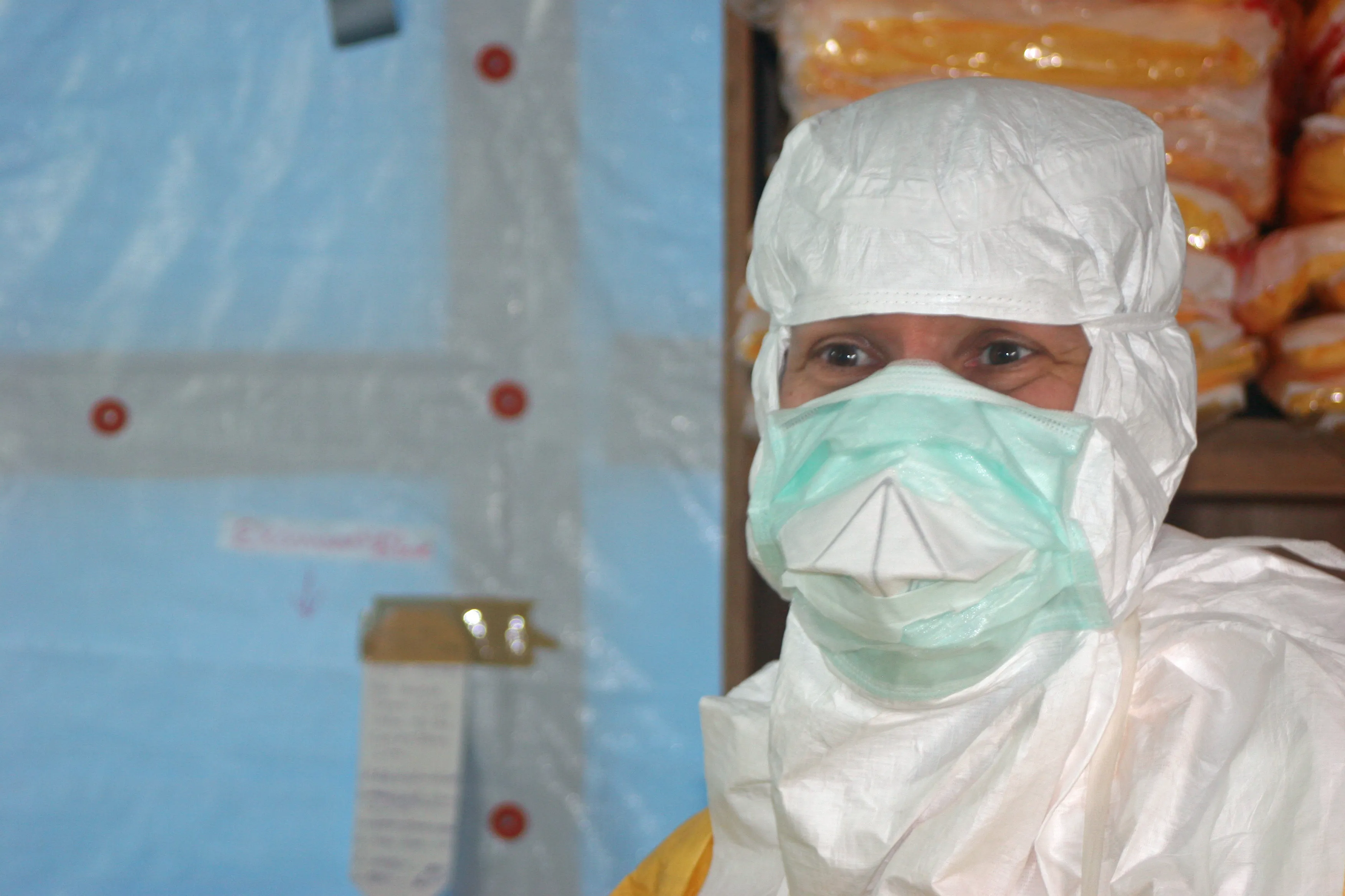 A public health official in a white protective suit, face shield, and blue nitrile gloves prepares to enter an Ebola treatment unit, shown from the side near the facility entrance