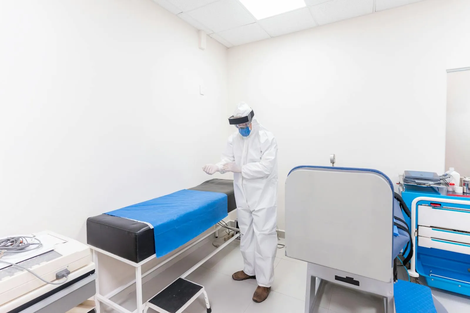 A health worker in a white protective coverall, face shield, mask and gloves stands in a clinical treatment room.