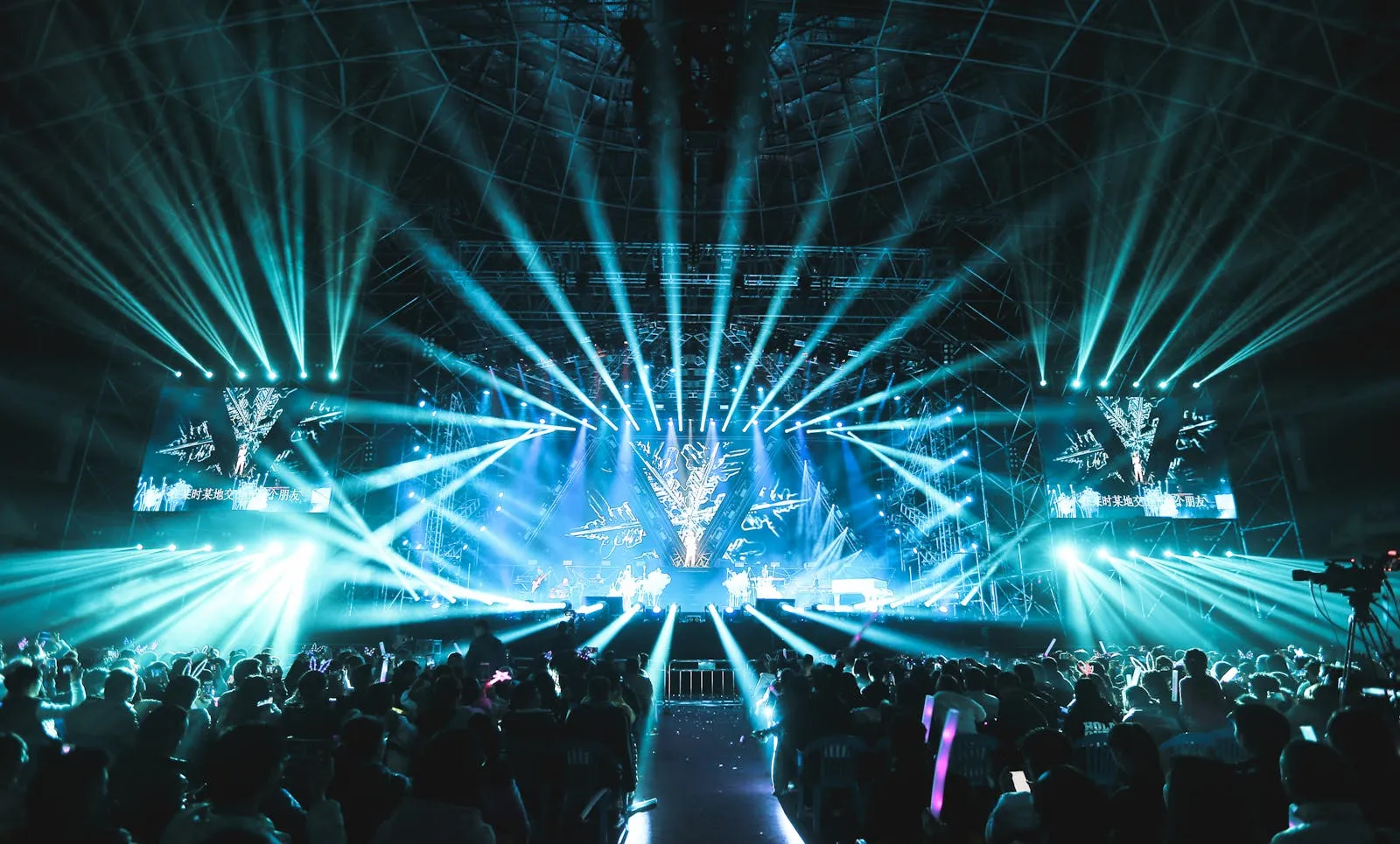 A wide concert arena view of stage lights and a crowd holding light sticks during a large indoor music event
