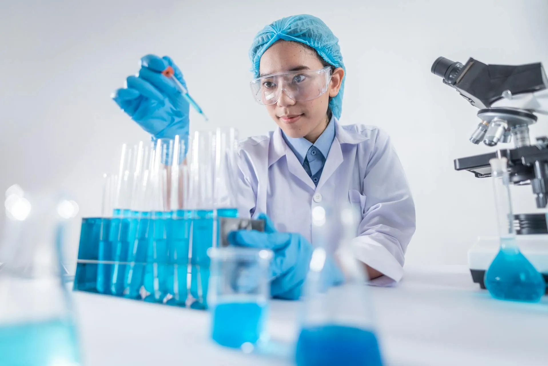 A researcher in a lab coat and safety goggles works with test tubes and a microscope in a bright laboratory