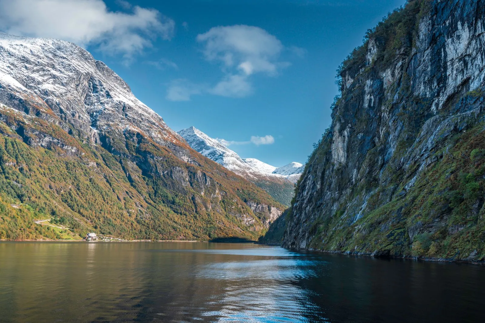 Rocky cliffs and snow-capped peaks reflected in the still dark-green waters of a Norwegian fjord on an autumn day