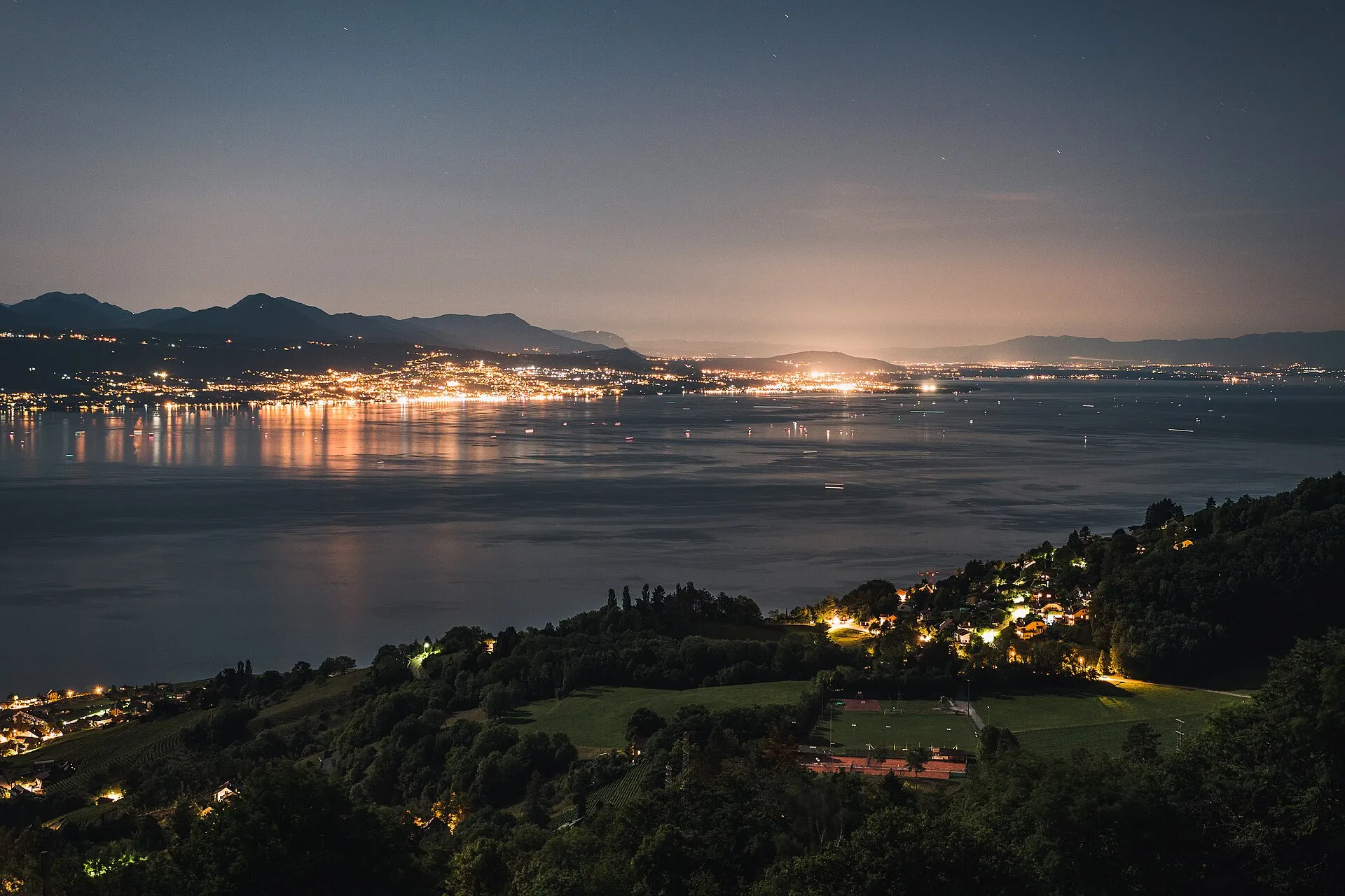 The lights of Évian-les-Bains glowing along the shore of Lake Geneva at night, seen across the water from the hills above.