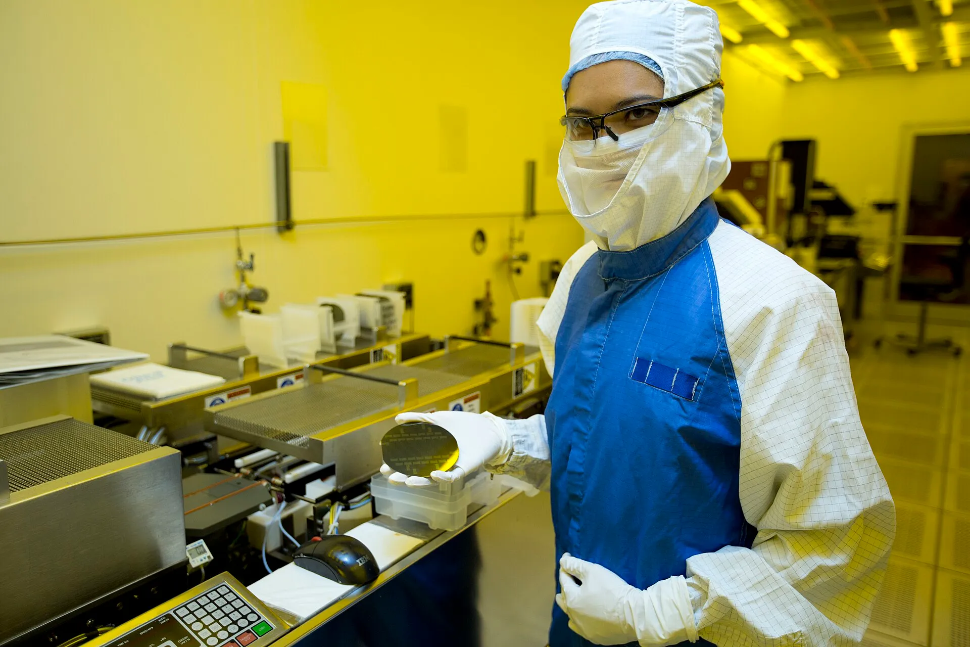 A gloved researcher holds a circular silicon wafer up to the light in a laboratory clean-room setting.