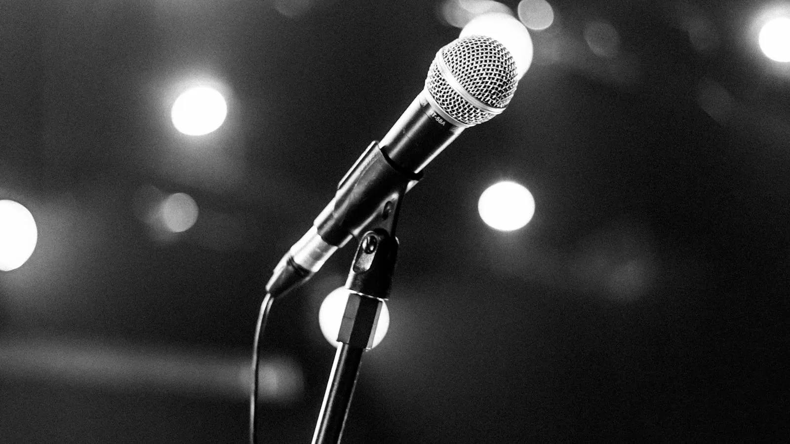 Black-and-white photo of a stand-up microphone on a stand under blurred stage spotlights.