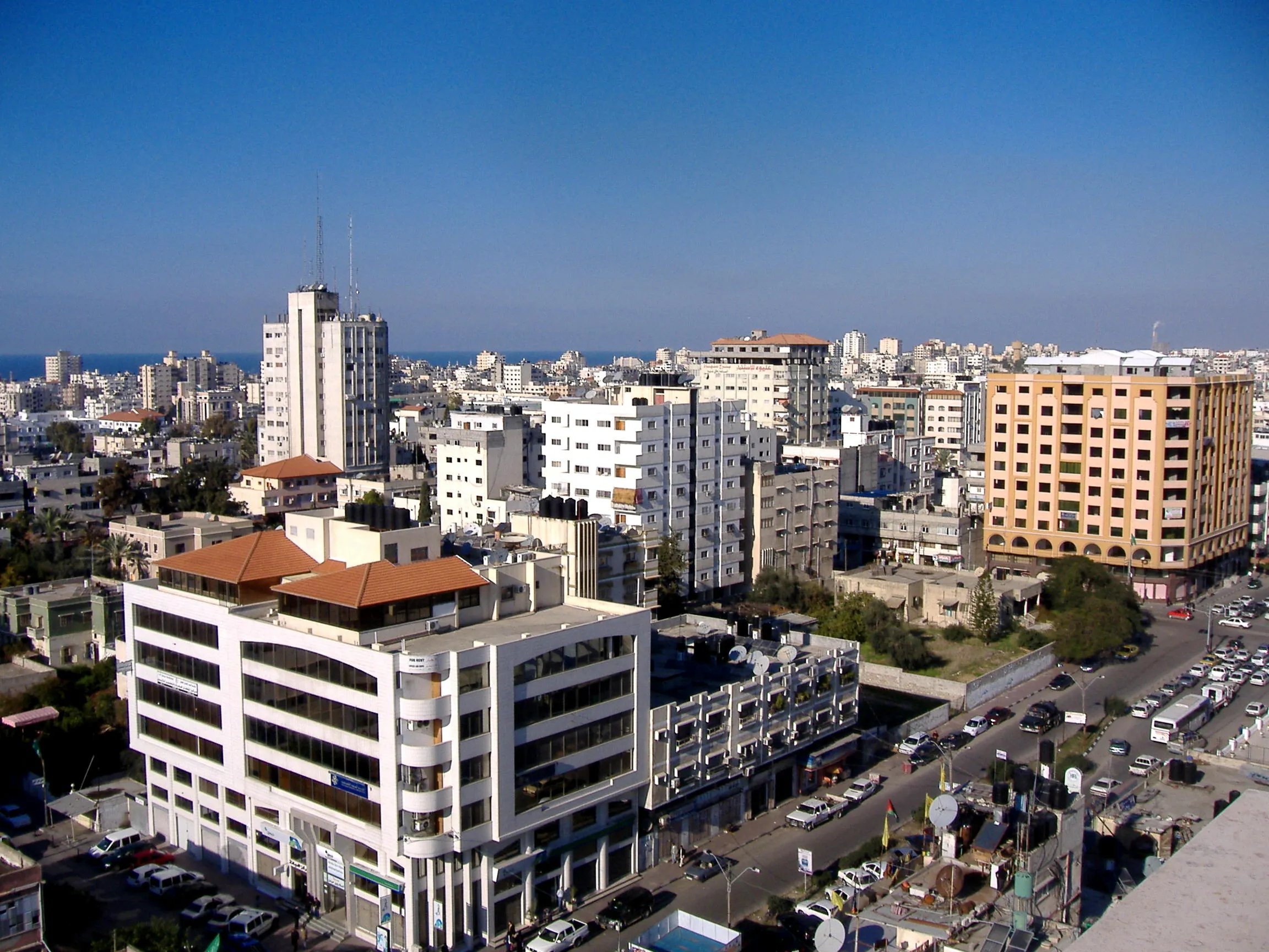 Elevated view of Gaza City with densely packed multi-story residential and commercial buildings extending toward the Mediterranean coastline under a clear blue sky