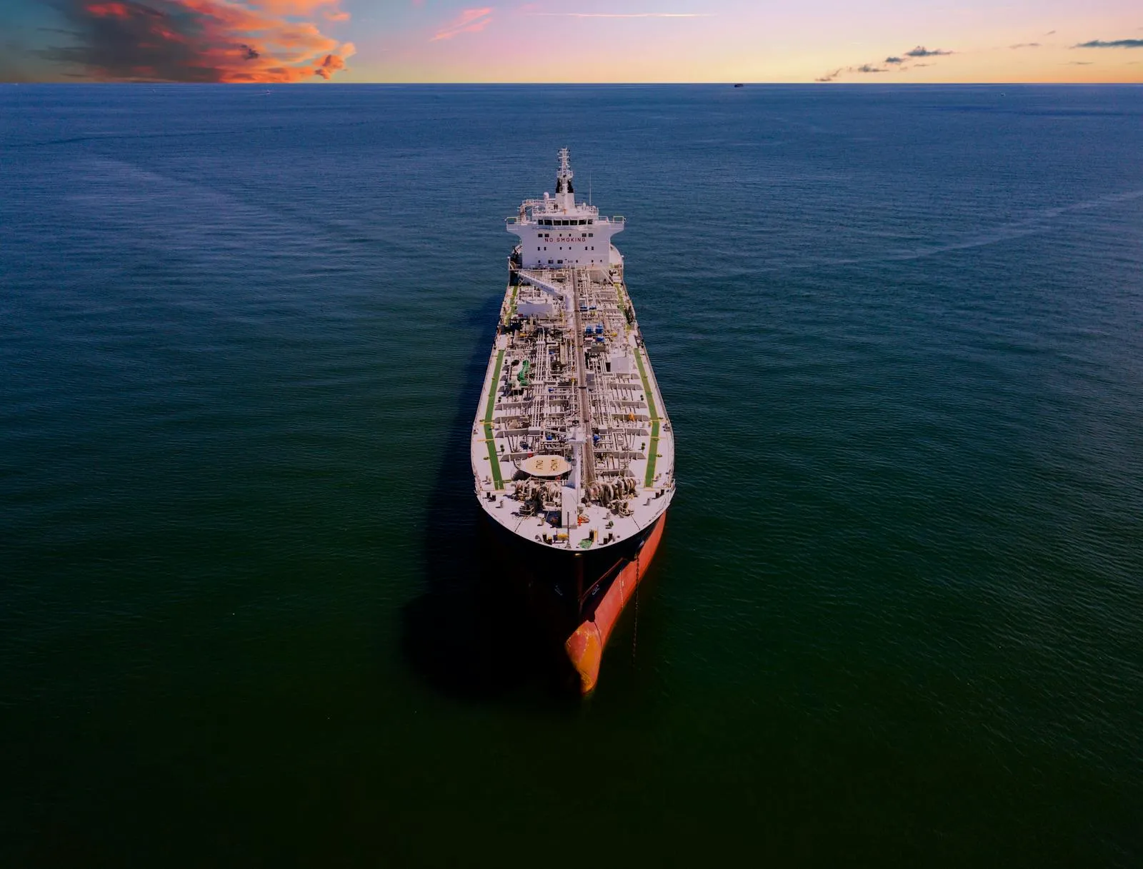 Aerial drone view of a large crude oil tanker navigating open ocean, seen from above at the bow, against a sunset-lit horizon