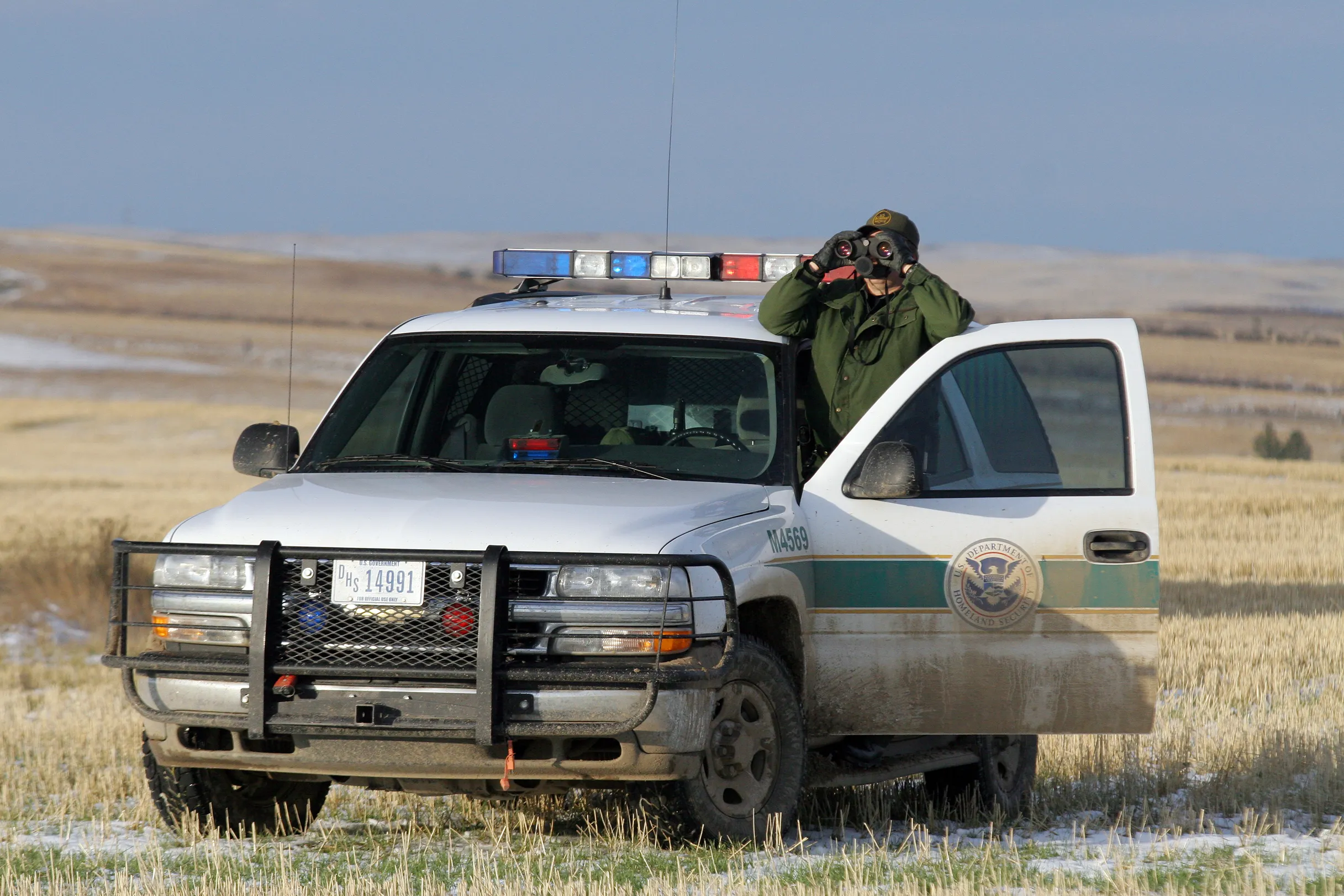 A U.S. Border Patrol agent in uniform stands beside a white Chevrolet Tahoe SUV holding binoculars on open terrain.