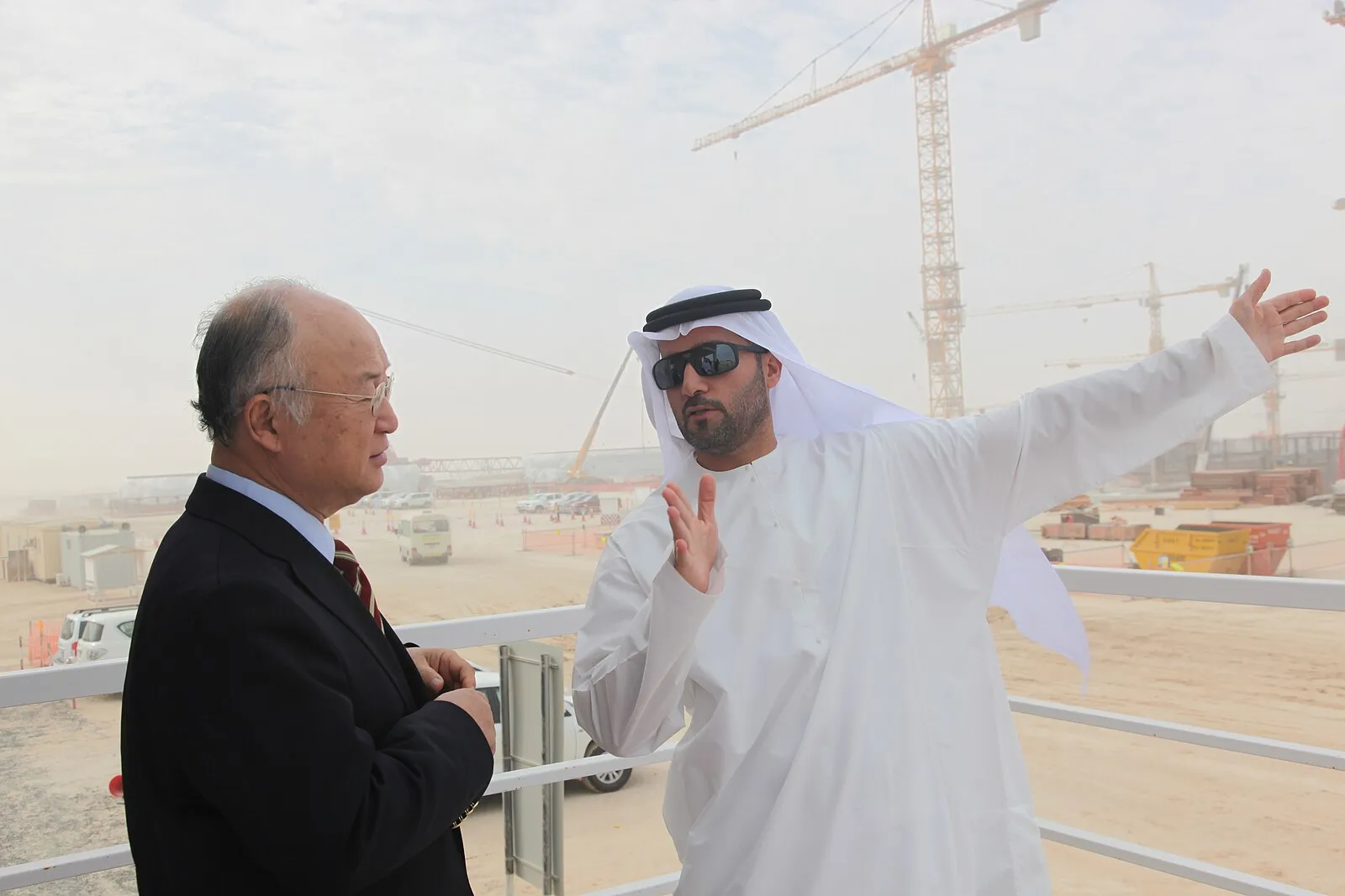 An official in hard hat and safety vest at the Barakah nuclear plant, with reactor containment domes visible behind.