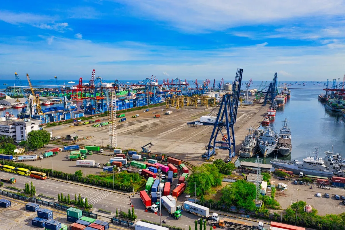 Aerial view of a busy international port with cargo ships moored alongside docks lined with container cranes and shipping infrastructure under a clear blue sky.