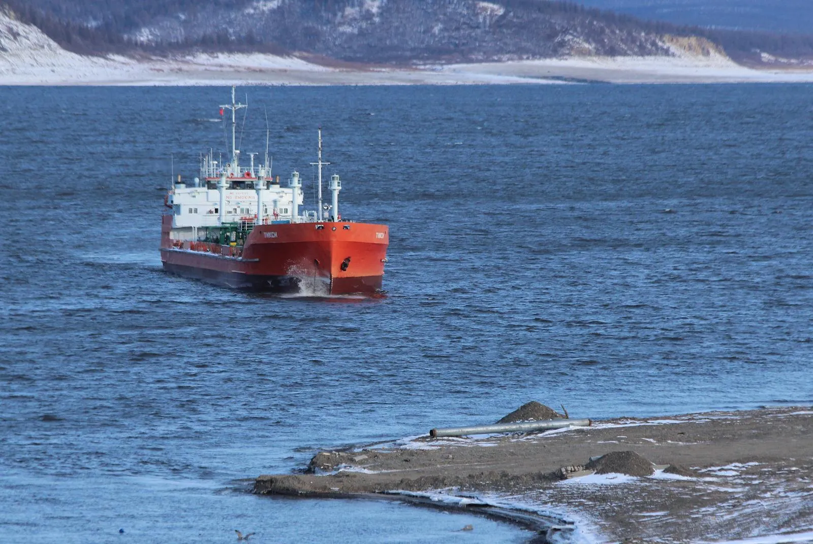 An orange oil tanker ship navigating a wide river channel with snow-dusted hills visible along the far bank.