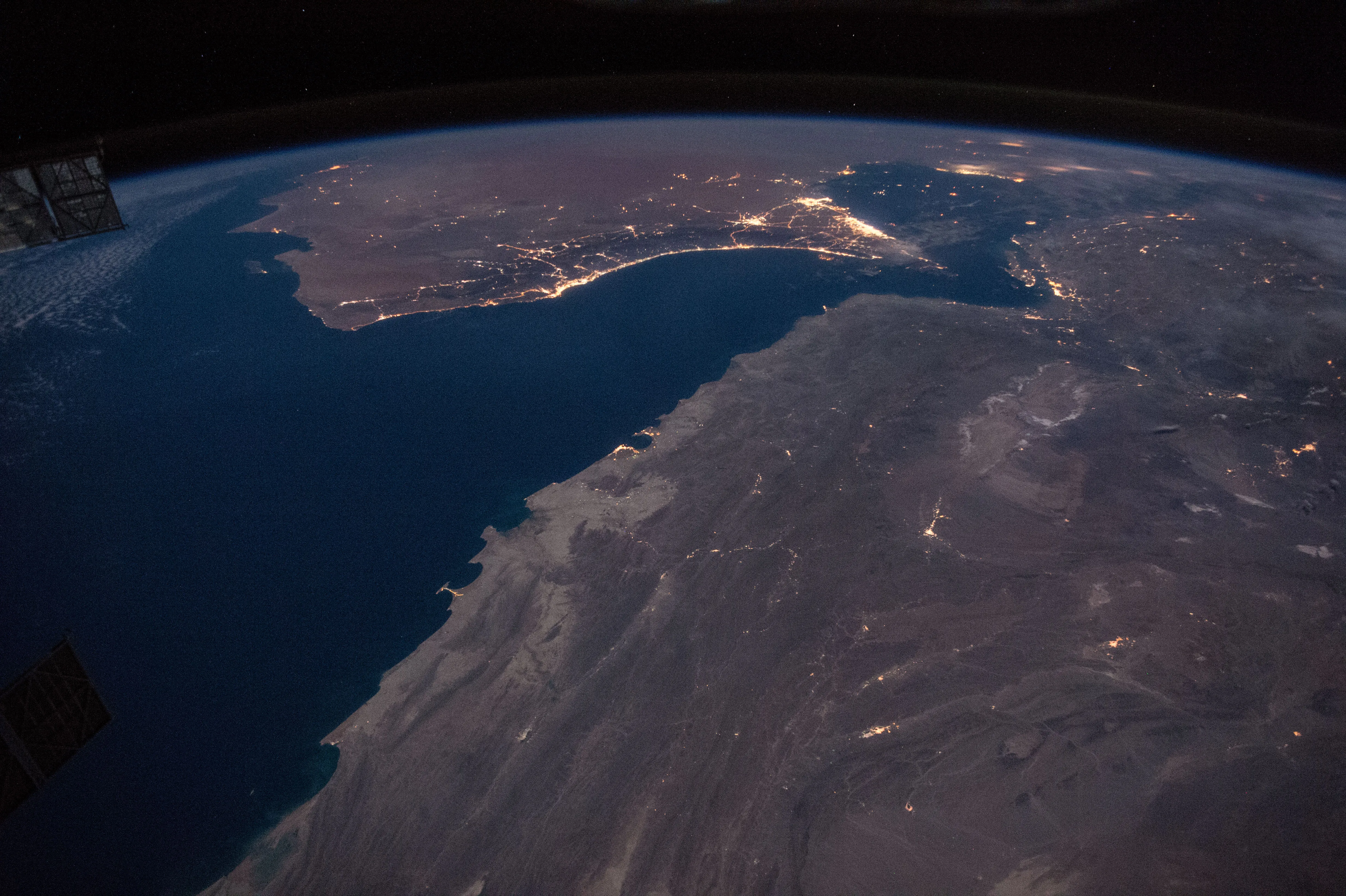 Nighttime aerial view from the International Space Station showing city lights along the Gulf of Oman coastline, with the dark water of the Persian Gulf and the Strait of Hormuz at center