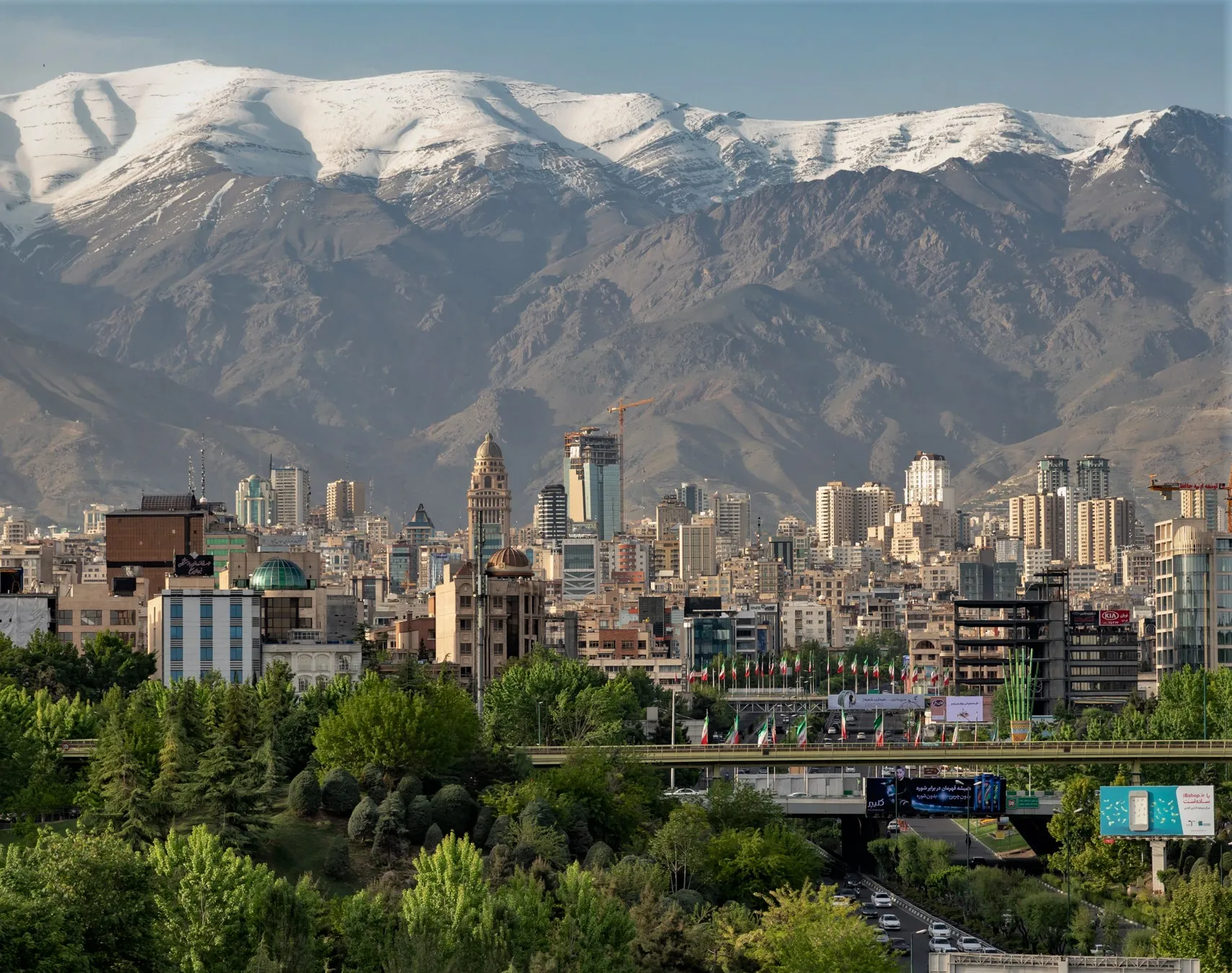 Tehran city skyline viewed from near the Tabiat Bridge, with apartment towers and commercial buildings stretching toward Tochal mountain under a hazy sky