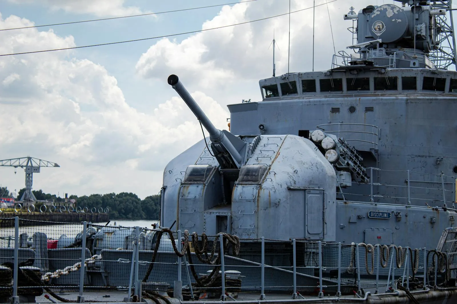 A gray naval warship with a forward gun turret moored at an industrial harbor, cranes and a green shoreline visible in the background