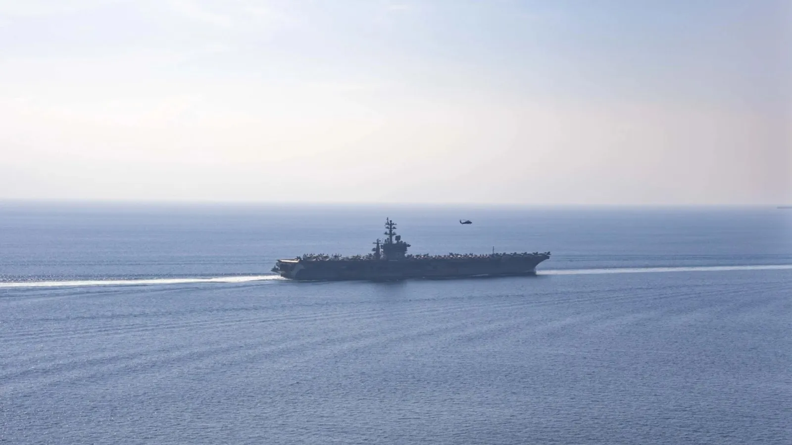 The aircraft carrier USS Dwight D. Eisenhower transiting calm water in the Strait of Hormuz, viewed from above, with a helicopter overhead.