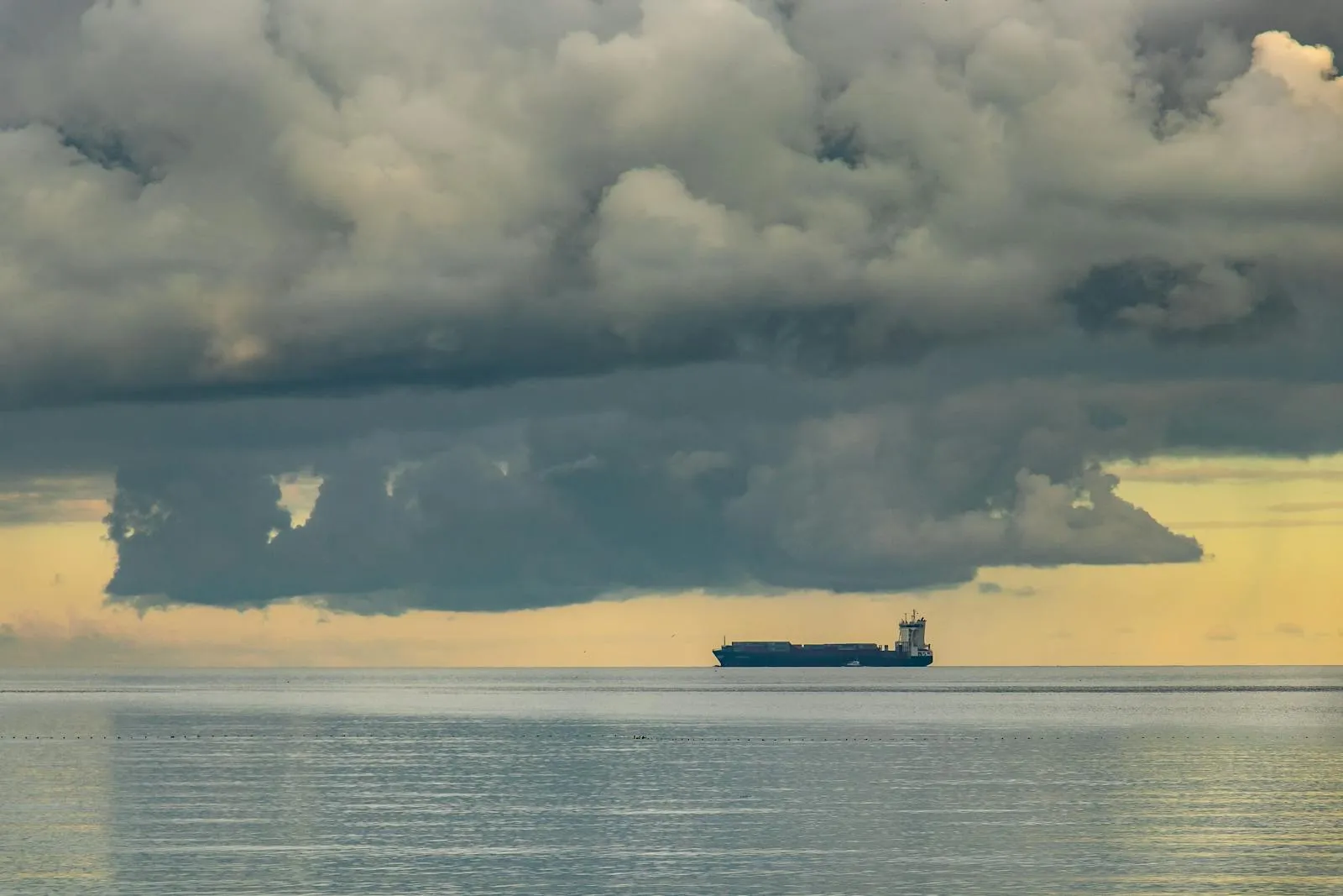 A large cargo ship sits on the horizon under dramatic dark storm clouds over a calm sea