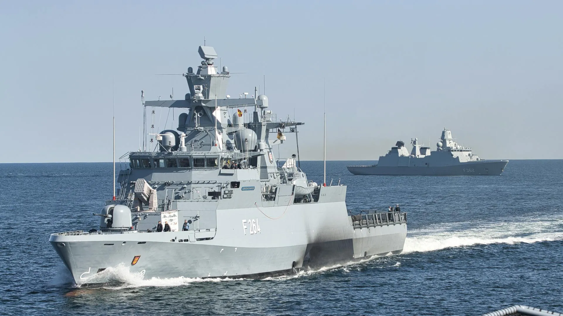 Two gray naval warships underway on calm open sea under a pale blue sky, the closer vessel showing its port side as it cuts a white wake.
