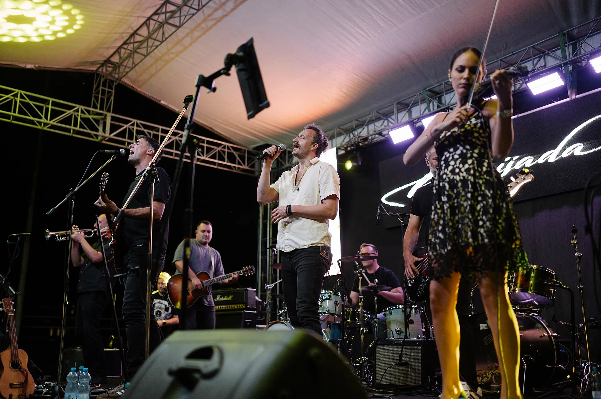 A band performs on a brightly lit outdoor festival stage at night as a large crowd watches.