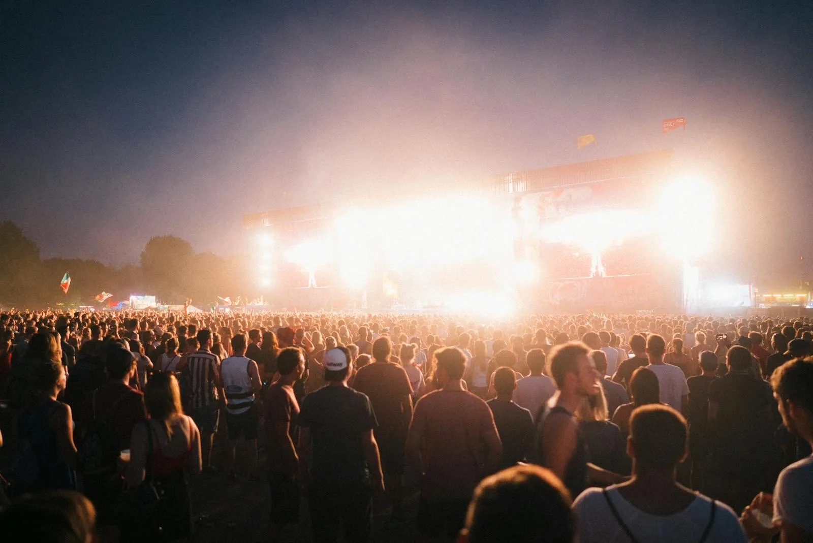 A massive outdoor music festival crowd at night facing a brightly lit main stage with giant LED screens