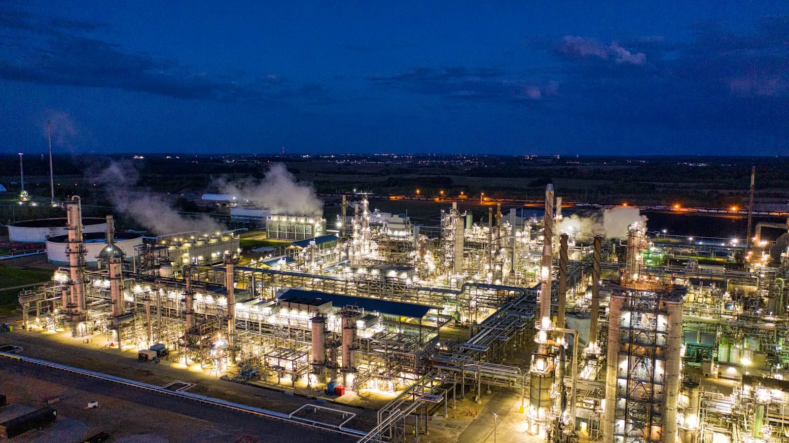 Aerial nighttime view of a brightly lit oil refinery complex with industrial flare stacks and an extensive network of pipes glowing against the dark surroundings