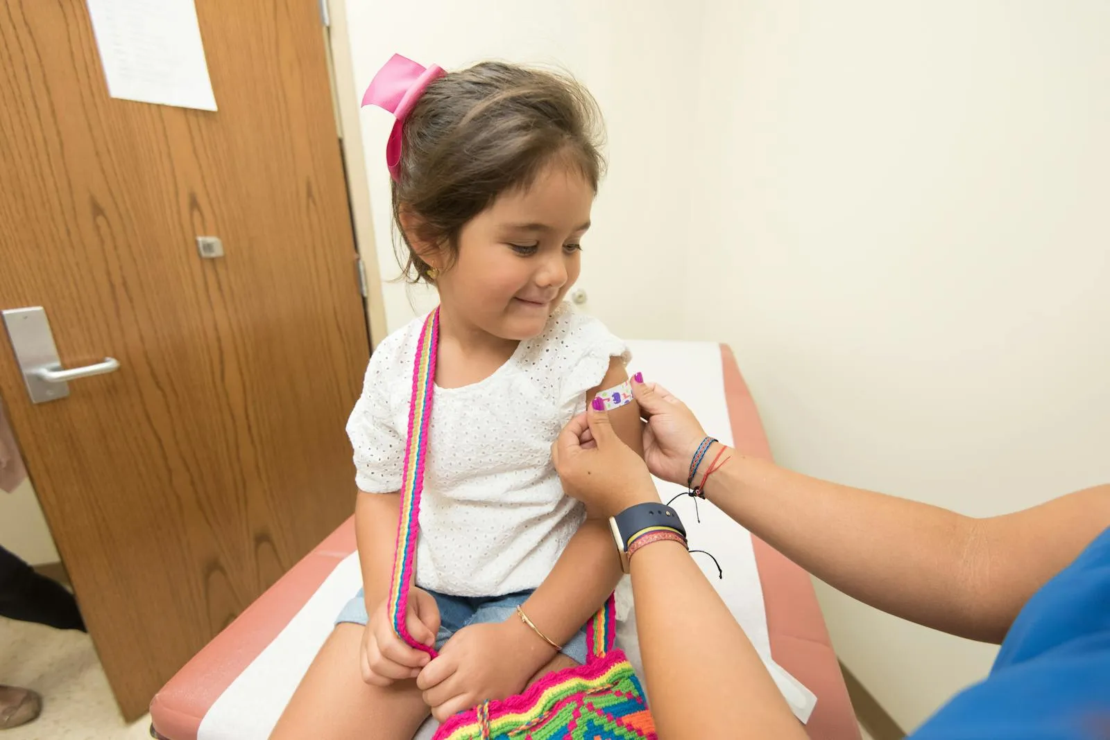 A young girl smiling as a healthcare worker applies a bandage to her upper arm in a clinic exam room after receiving a vaccination
