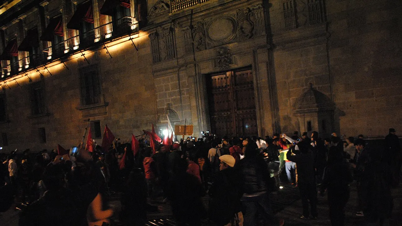 Protesters carrying red flags gather at night outside the lit facade of the National Palace in Mexico City's Zócalo