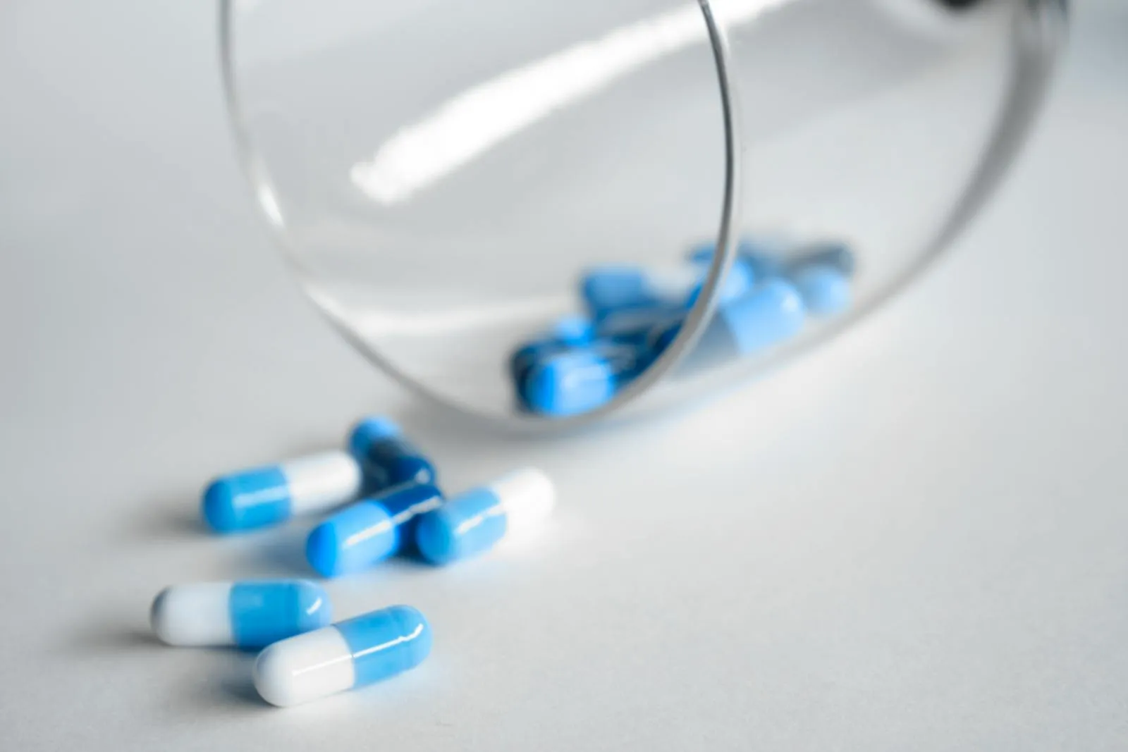 Blue and white prescription capsules spilling from an overturned clear medicine bottle onto a white surface