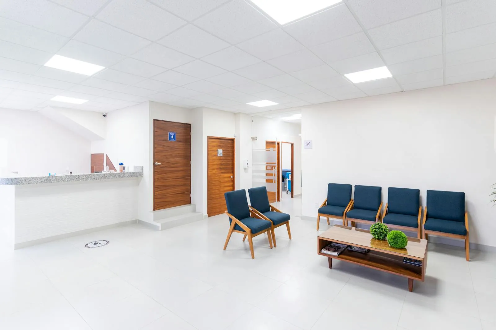An empty medical waiting room with blue upholstered chairs, a reception desk, and wooden doors.