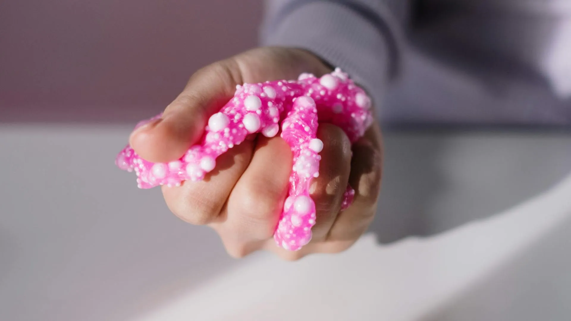 A close-up of a hand squeezing a piece of bright pink slime filled with small white foam beads.