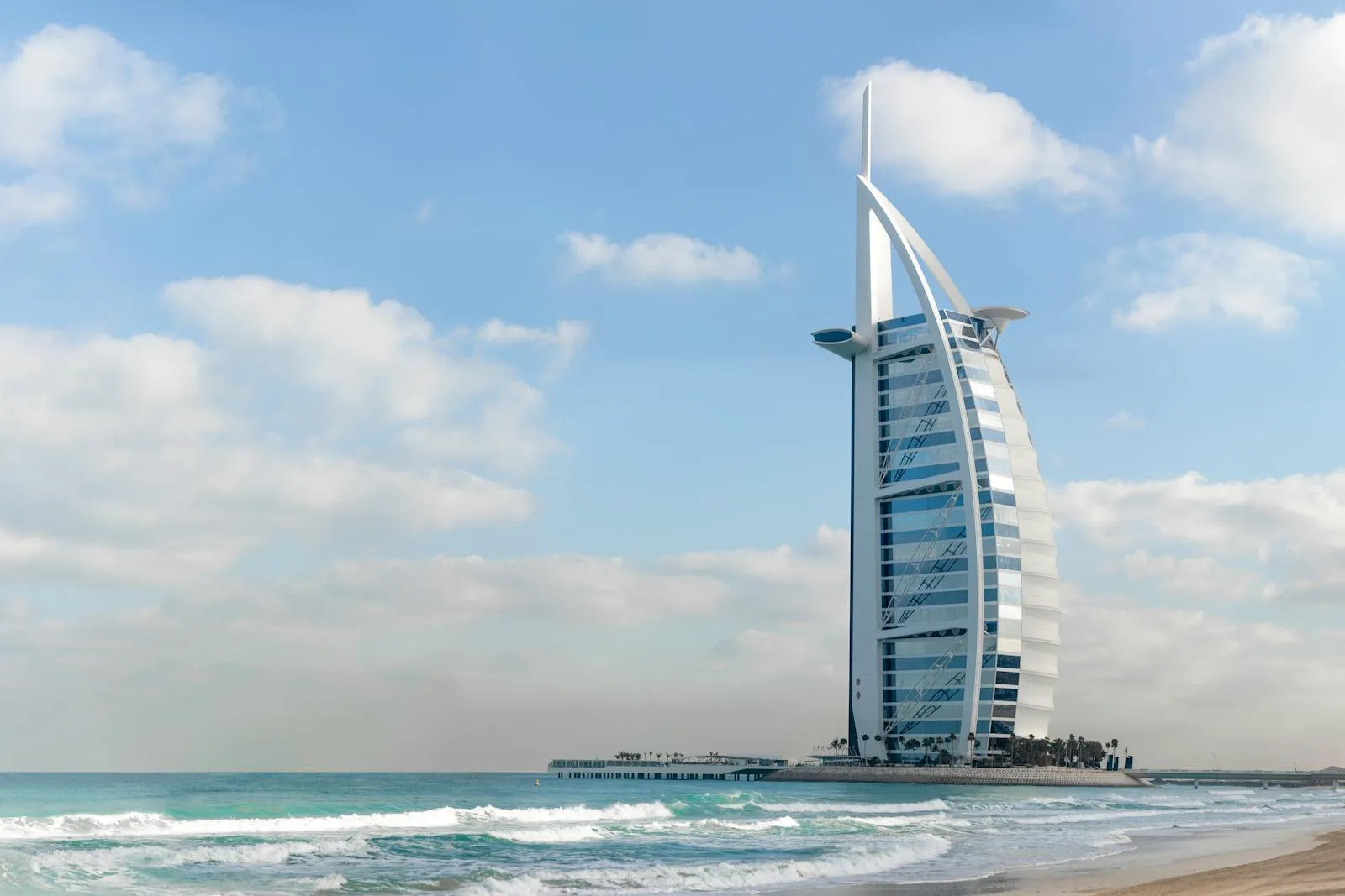 The Burj Al Arab tower in Dubai, United Arab Emirates, photographed from the beach under a partly cloudy sky, with waves breaking on the sand in the foreground.