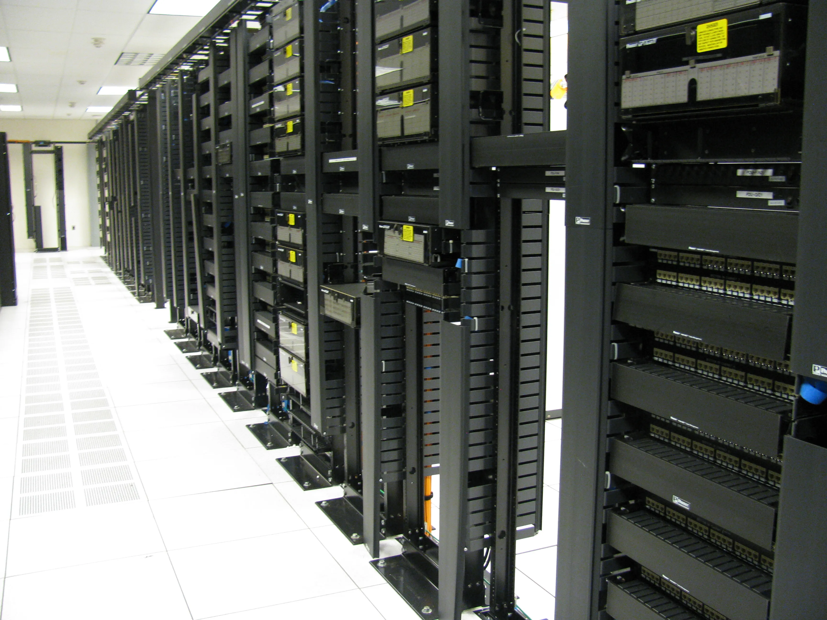 The front aisle of a large data center, showing rows of network switches and server equipment mounted in metal racks under fluorescent lighting.