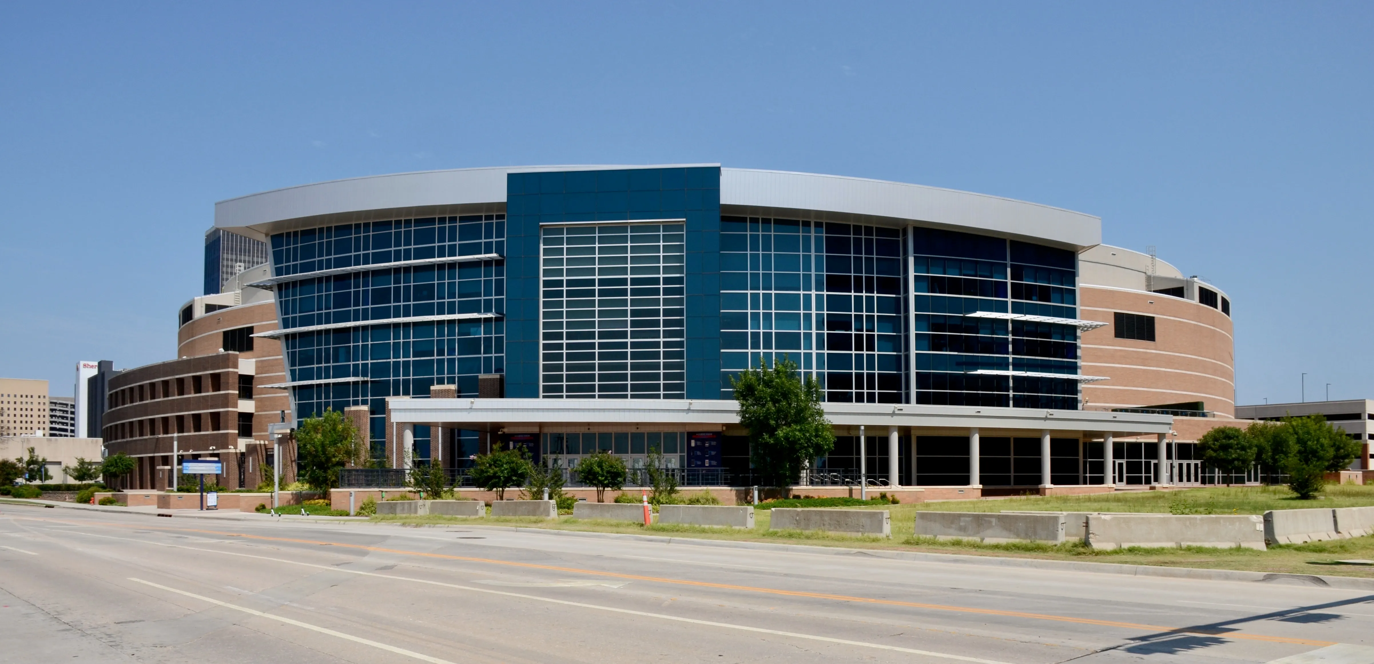 Paycom Center arena in Oklahoma City, a large modern building with blue glass and brick exterior, photographed from across the street under a clear blue sky