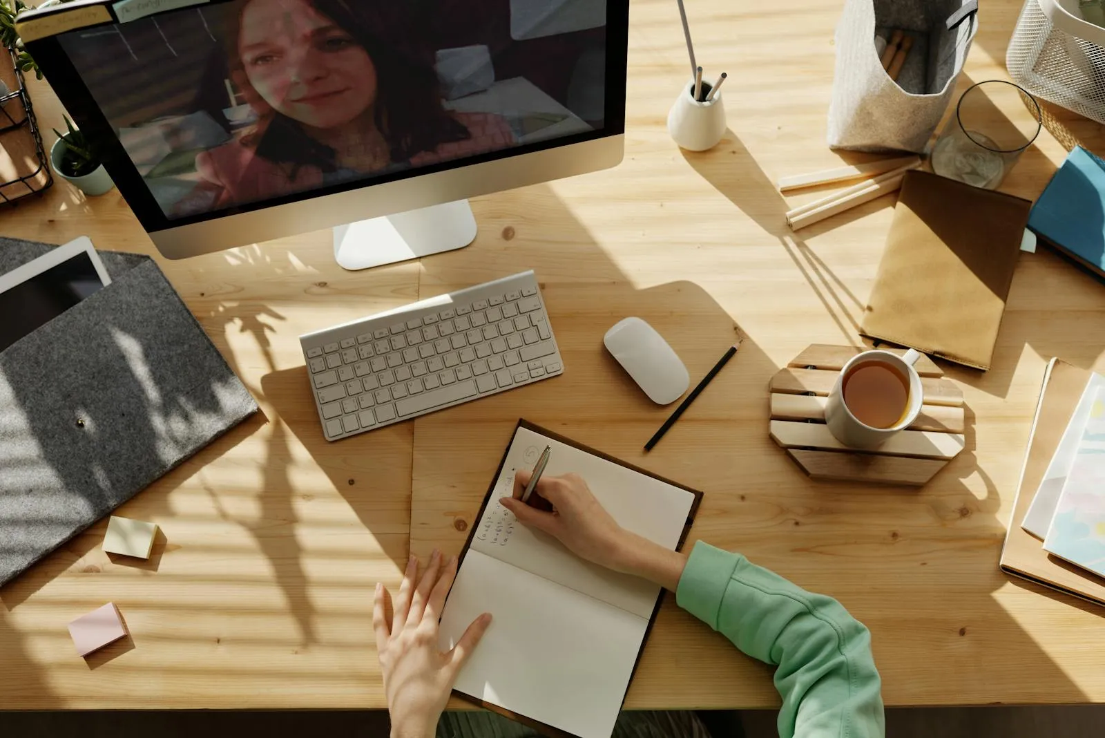 Top-down view of a wooden desk with an iMac showing a video call on screen, beside a keyboard, mouse, and open notepad.