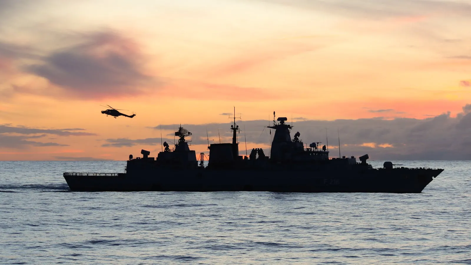 A dark silhouette of a naval warship moving across open sea waters against a pale gray overcast horizon.