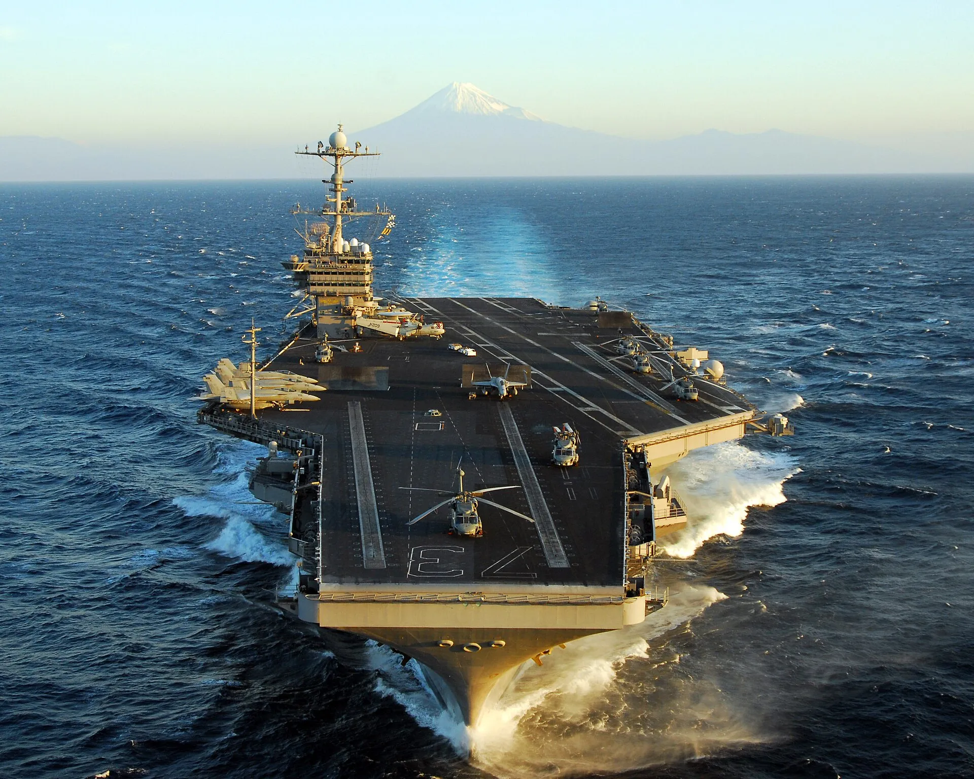 A U.S. Navy aircraft carrier steams through the Pacific Ocean with the snow-capped peak of Mount Fuji rising in the distance.