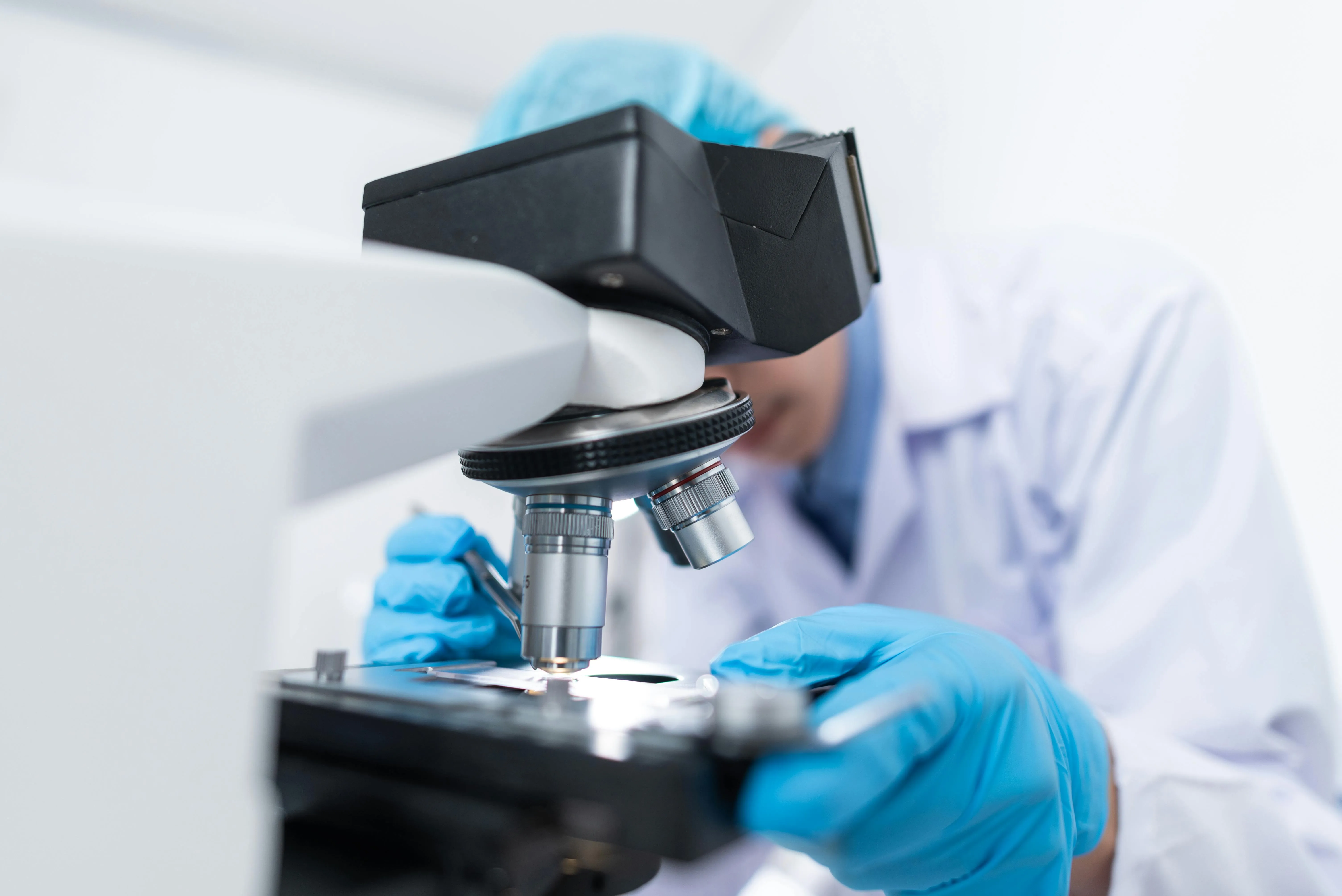 Researcher in a white lab coat and blue gloves adjusting a slide on a compound microscope in a laboratory