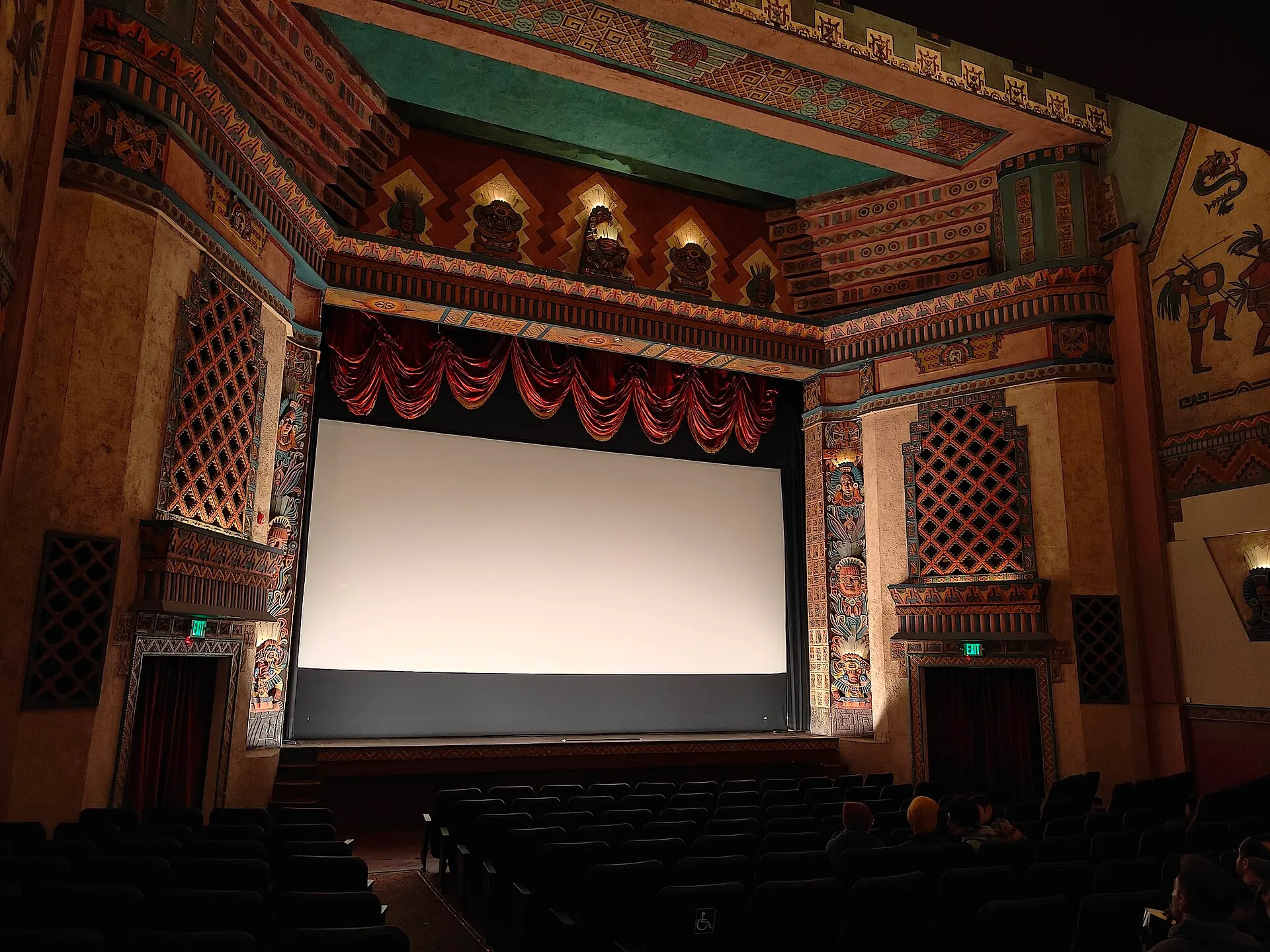 An ornate movie theater auditorium with a decorated proscenium, red curtains, a blank screen and empty seats.
