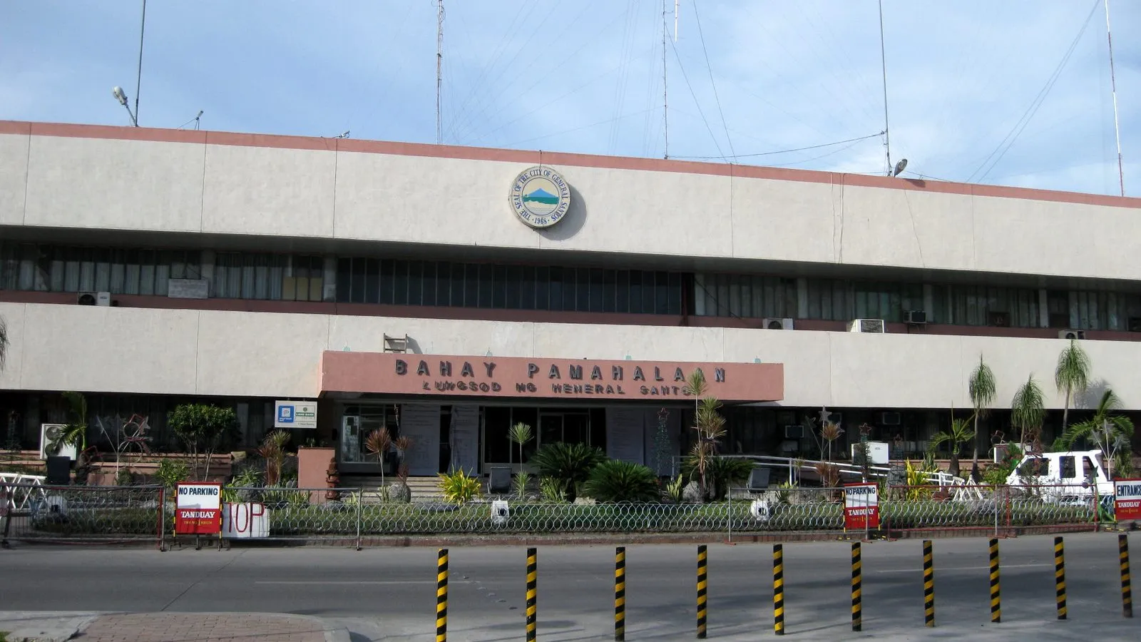 A modern multi-story city hall building with a curved facade and rows of flagpoles out front, under a clear blue sky in General Santos City, Philippines.