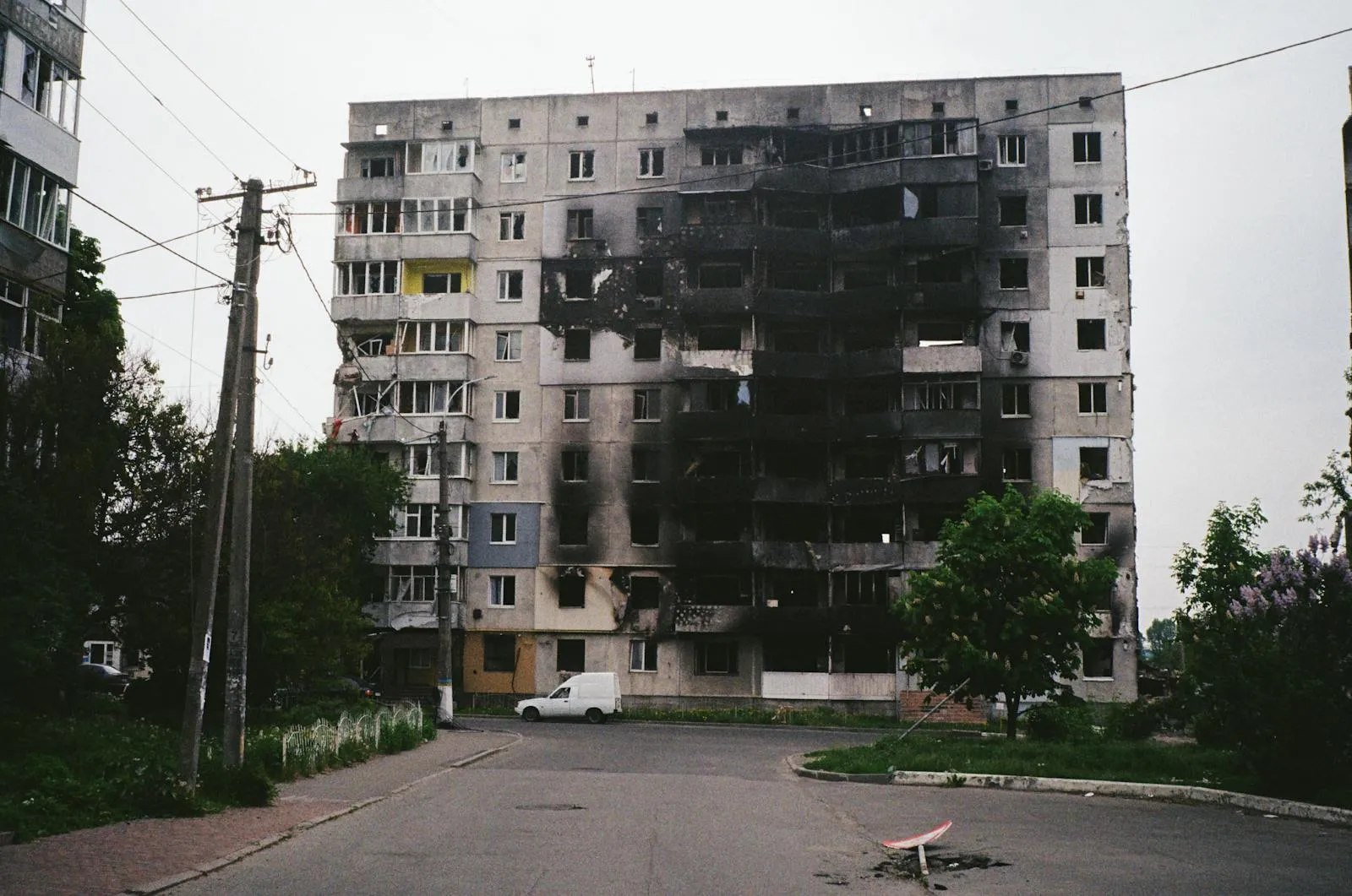 A multi-story Soviet-era residential building showing severe fire and blast damage, with scorched walls and blown-out windows on a tree-lined street