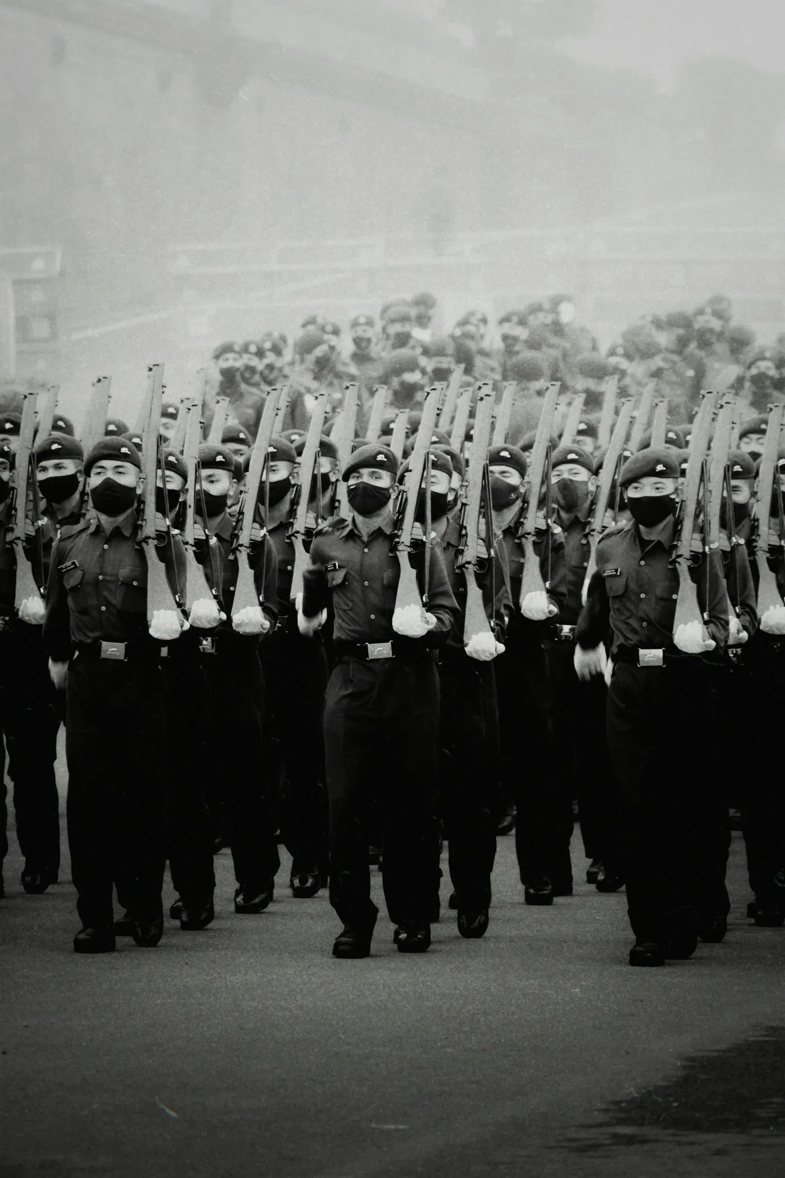Soldiers in dark dress uniforms march in tight formation carrying rifles during a military parade, photographed in black and white.