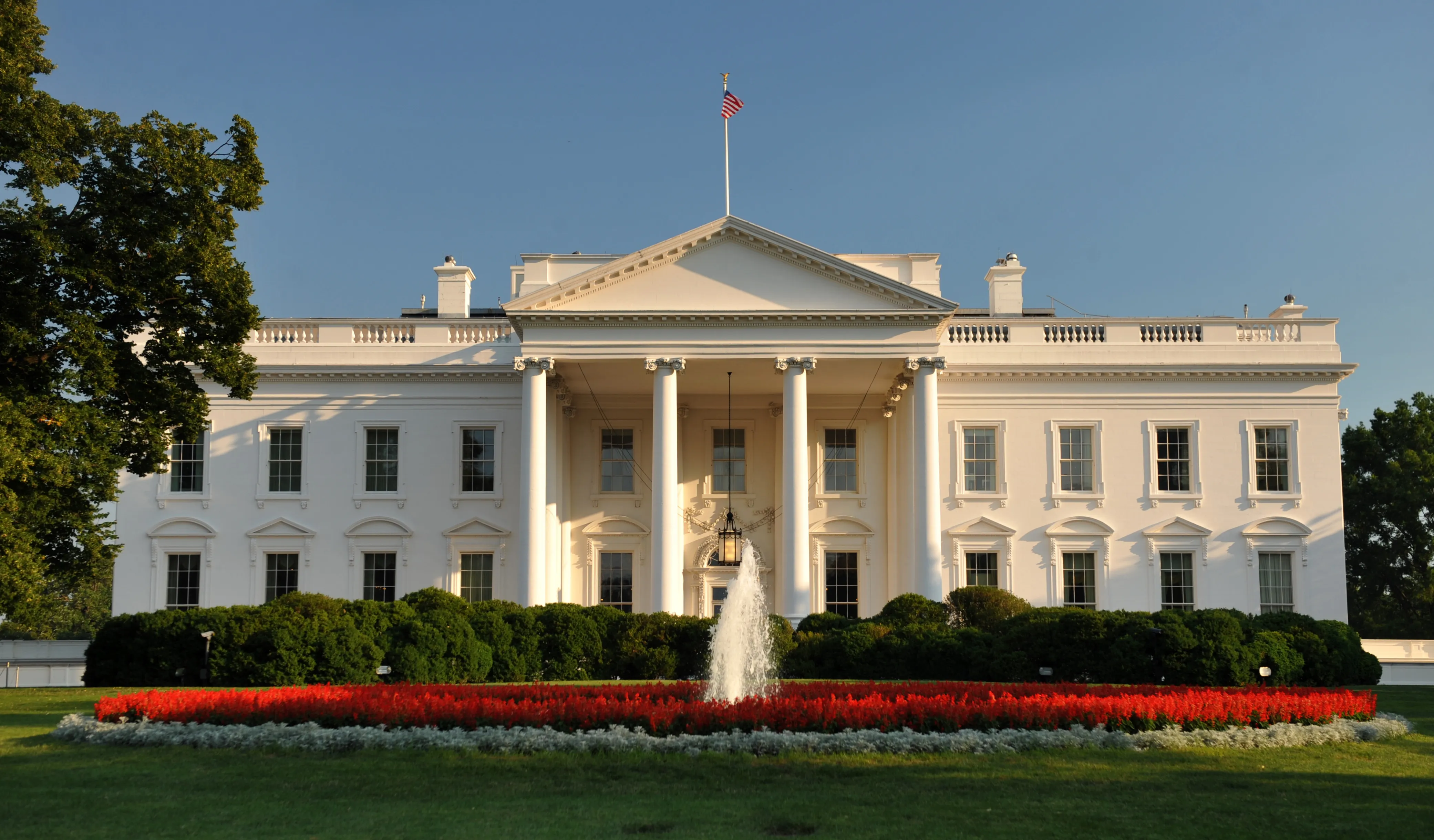 The north facade of the White House seen through the South Lawn, with the neoclassical portico and central flag under a clear blue sky